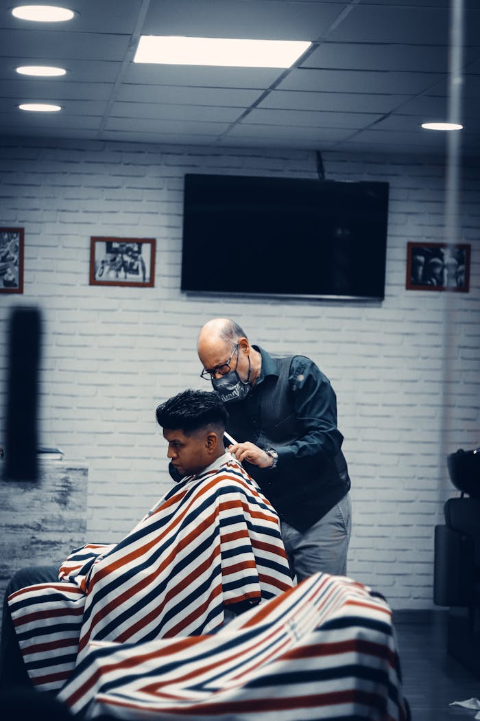 Senior barber in mask grooming young man in a modern indoor barbershop.